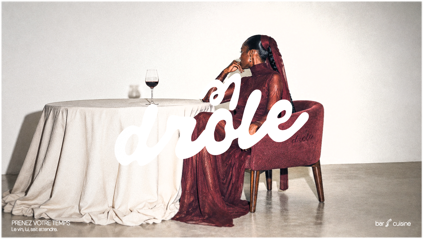 Drôle — mulher de vestido bordô sentada à mesa com taça de vinho e o logotipo manuscrito