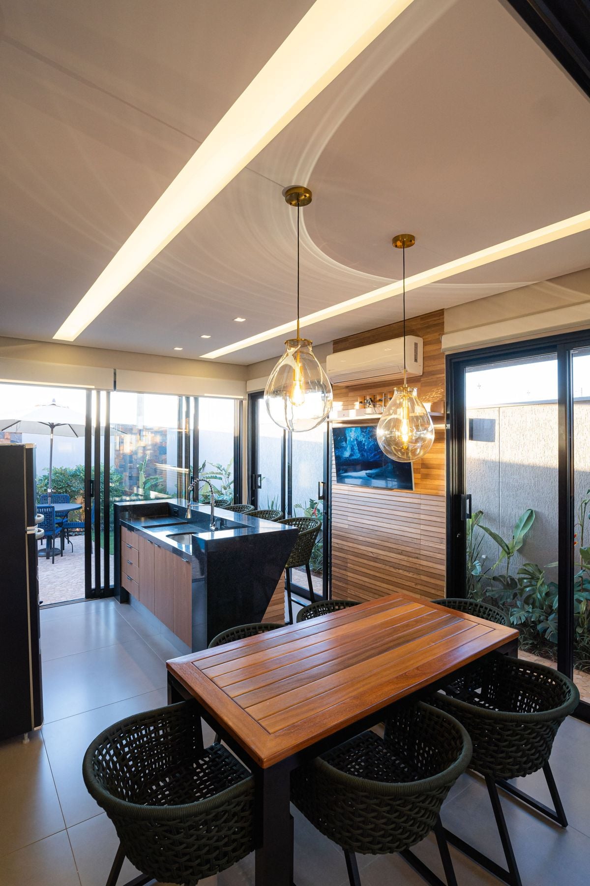 Área gourmet com mesa de madeira, ilha em granito preto e pendentes de vidro, projeto Quinta dos Ventos